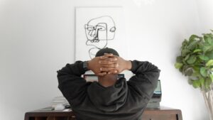 Young black man seated at desk with hands clasped behind his head with abstrack line drawing on wall in front of him. 